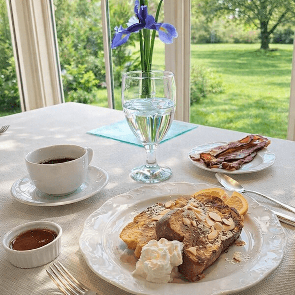 A table with coffee, french toast, bacon, and a glass of water in front of a sunny field