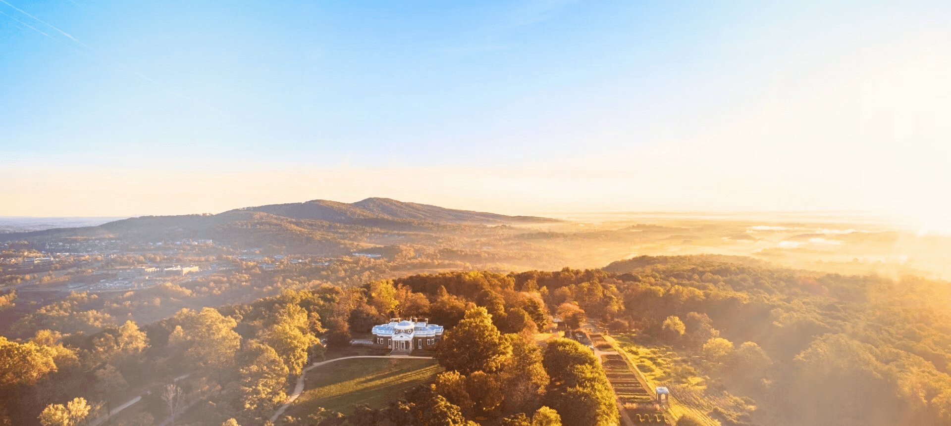 Aerial view of a lush landscape bathed in golden sunlight, featuring rolling hills and a white building surrounded by trees.